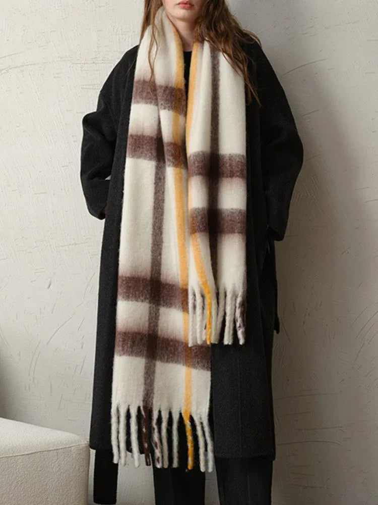 Full body shot of a woman wearing a long dark coat and a large plaid scarf with white, brown, and yellow stripes.