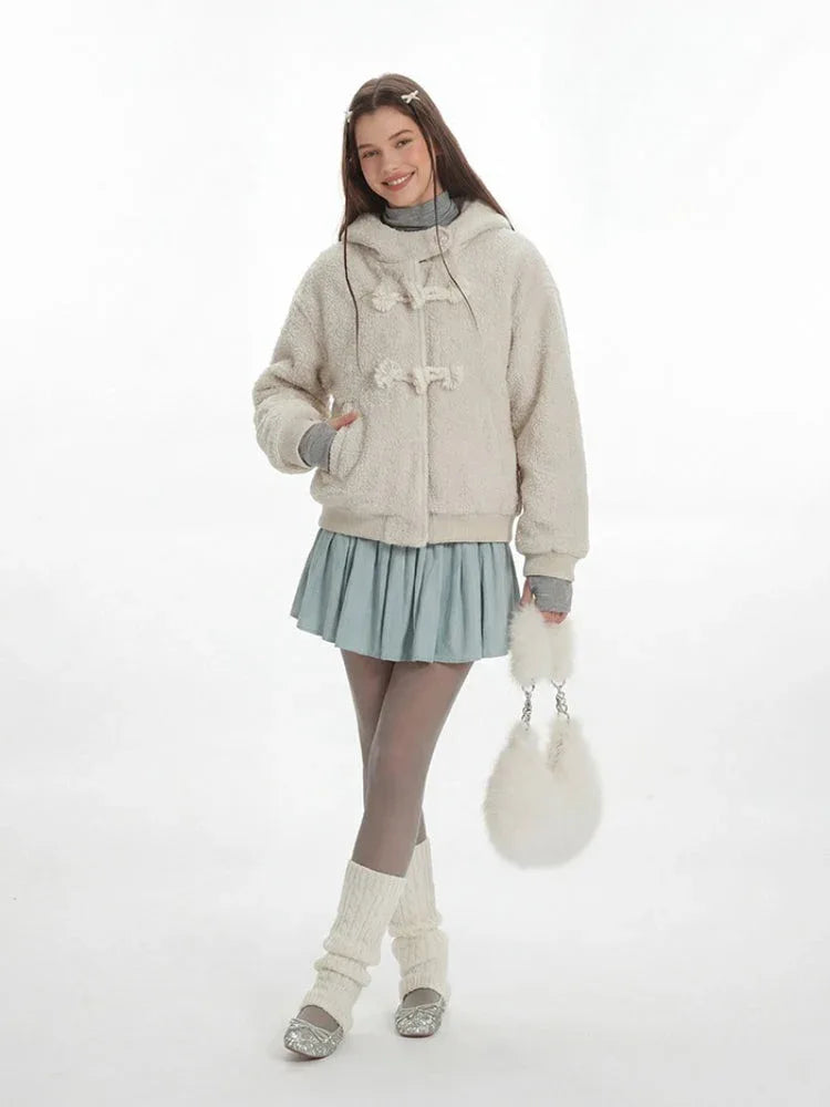 Model smiling and wearing a cream fuzzy hooded bomber jacket with toggle closure, a light blue pleated skirt, grey turtleneck, sheer tights, white leg warmers, and a white fuzzy handbag. Secondary image