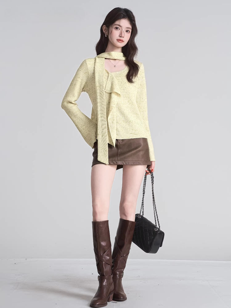 Full body shot of a woman wearing a light yellow textured knit top, brown mini skirt, and brown boots, holding a black handbag.