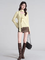 Full body shot of a woman wearing a light yellow textured knit top, brown mini skirt, and brown boots, holding a black handbag.