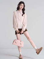 Full body shot of a woman wearing a light pink ruffled peplum blouse with a Peter Pan collar and lace trim, dark brown shorts, white socks, and pink sneakers, holding a pink handbag.
