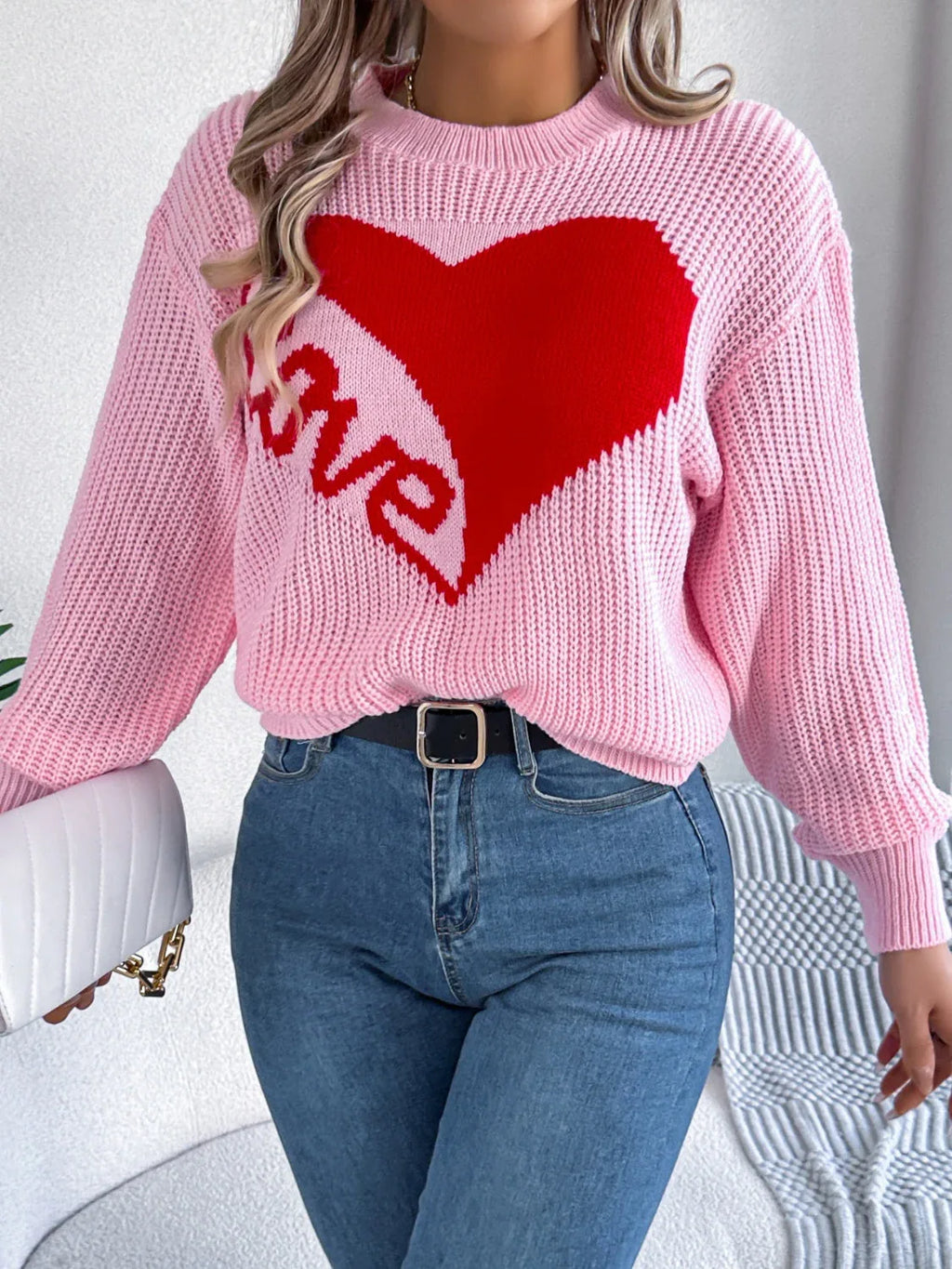 Woman wearing a pink knit sweater with a red heart graphic and the word "love" on the front, holding a white handbag.