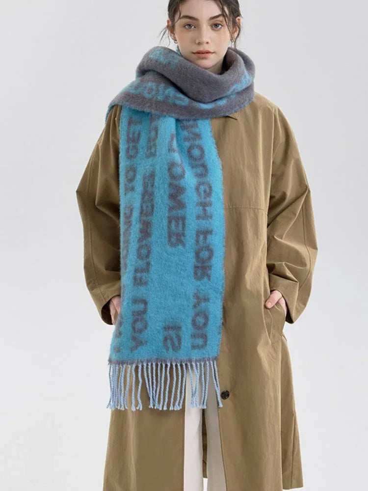 A woman models a blue and gray knit scarf with text print and fringe detail, worn over a tan trench coat.