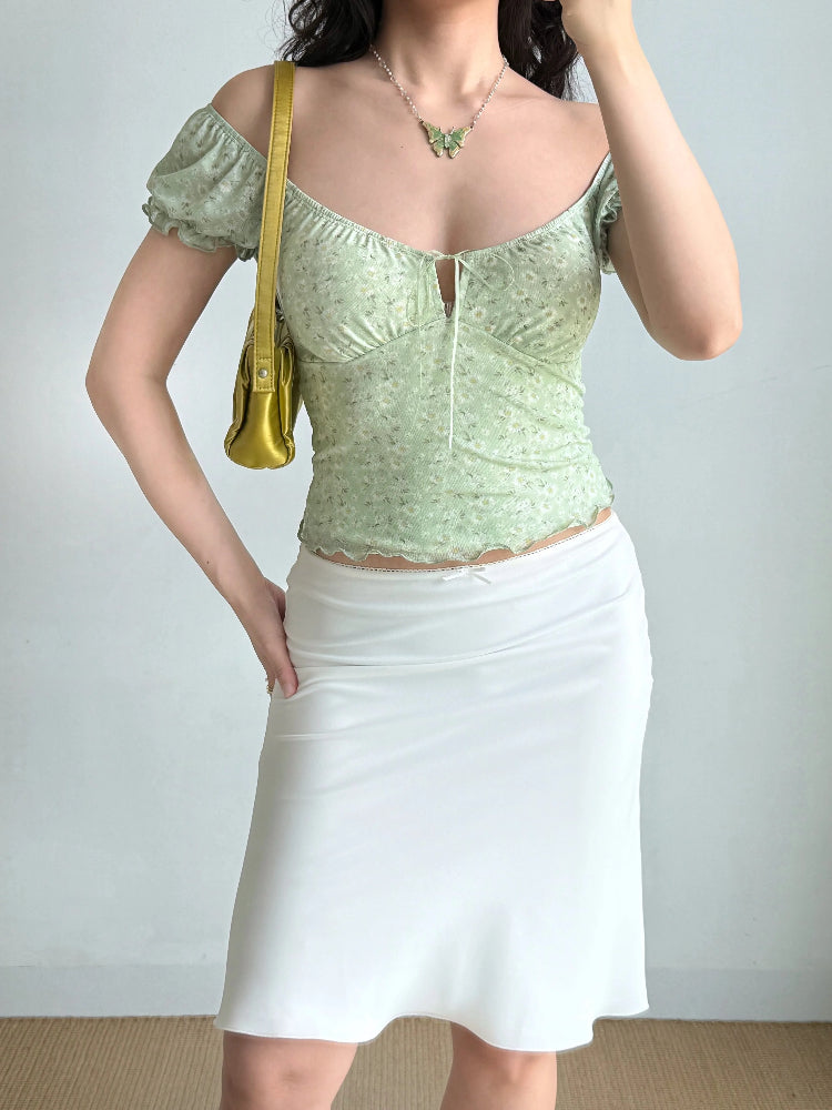 Full-body shot of a model wearing a light green floral print top, a white skirt, and a yellow handbag.