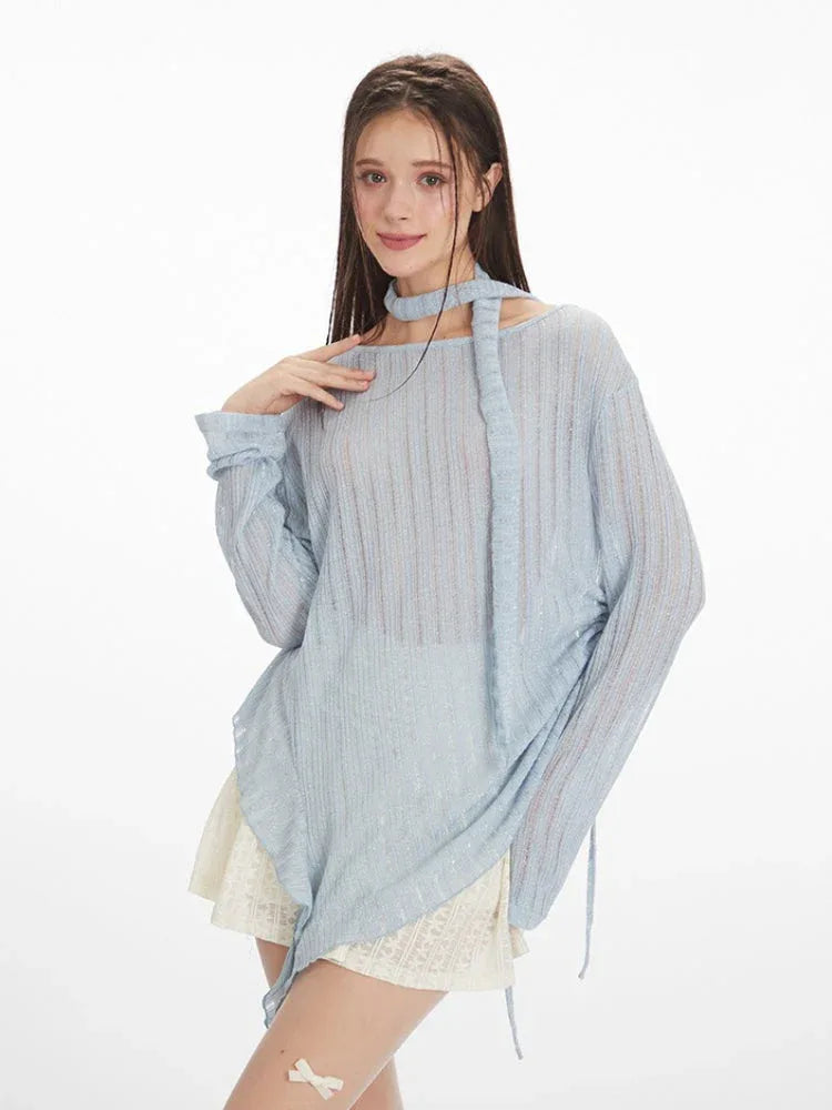 front view of a model wearing a light blue sheer knit sweater with an asymmetrical hem and a neck scarf, paired with a white lace skirt and white heels, holding a white handbag, posing with hand on chest