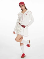 Model posing in a white knit sweater with a sailor collar and red trim, white pleated skirt, red belt bag, white socks, and red sneakers.