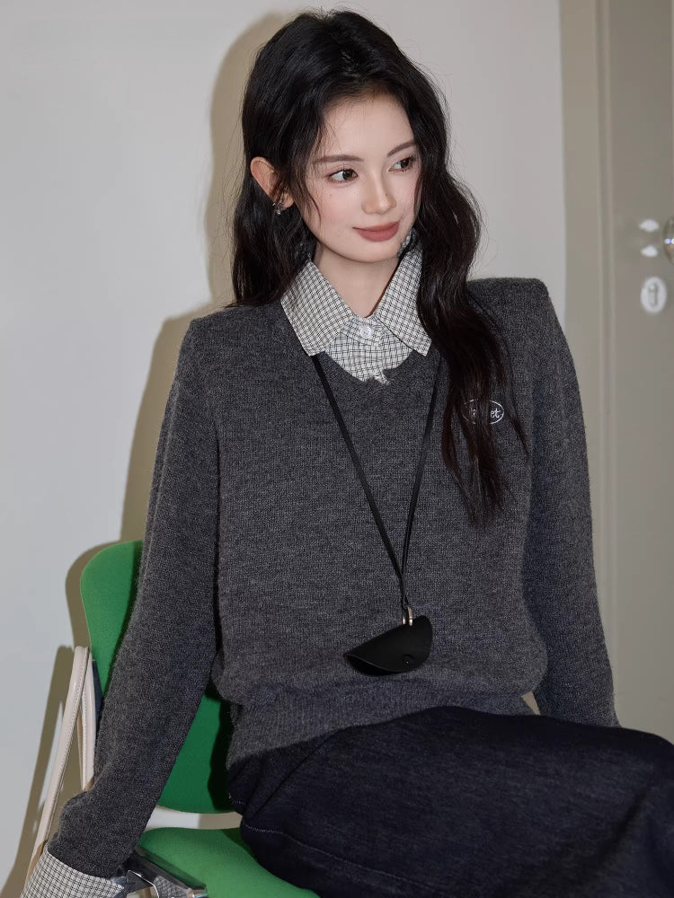 Woman sitting on a green chair wearing a grey v-neck sweater over a checkered collared shirt, dark wash wide leg jeans, and a black crossbody pouch.