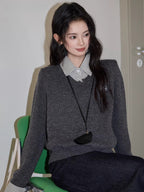 Woman sitting on a green chair wearing a grey v-neck sweater over a checkered collared shirt, dark wash wide leg jeans, and a black crossbody pouch.