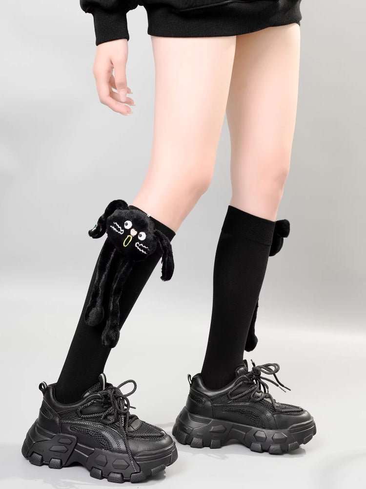 View of a person wearing knee-high black socks with a plush black cat charm on the left leg.