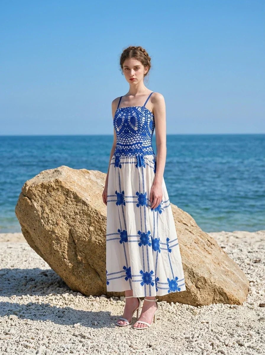 Woman wearing a blue and white crocheted and embroidered maxi dress standing on a beach near a large rock.
