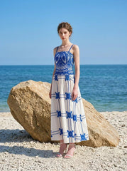 Woman wearing a blue and white crocheted and embroidered maxi dress standing on a beach near a large rock.