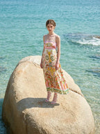 Woman wearing a white maxi dress with a colorful paisley print and pink smocked bodice, standing on a large rock in the ocean.