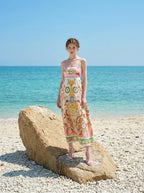 Woman wearing a white maxi dress with a colorful paisley print and pink smocked bodice, standing on a large rock on a beach with the ocean in the background.