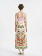 Rear view of a woman wearing a white maxi dress with a colorful paisley print and pink smocked bodice, standing against a white background.
