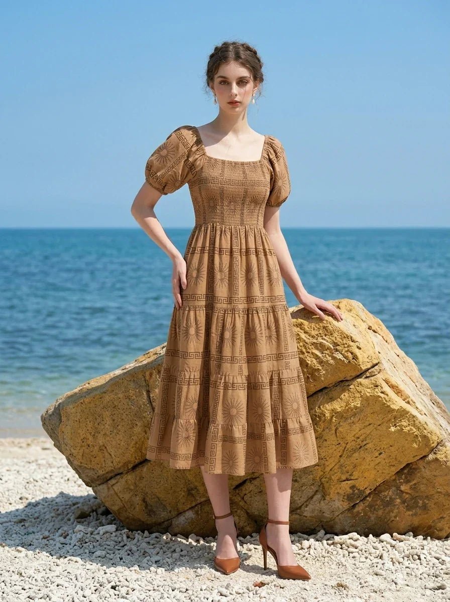 Woman wearing a brown puff-sleeve midi dress with a smocked bodice and tiered skirt featuring a sun and geometric print, posing on a beach next to a large rock with the ocean in the background.