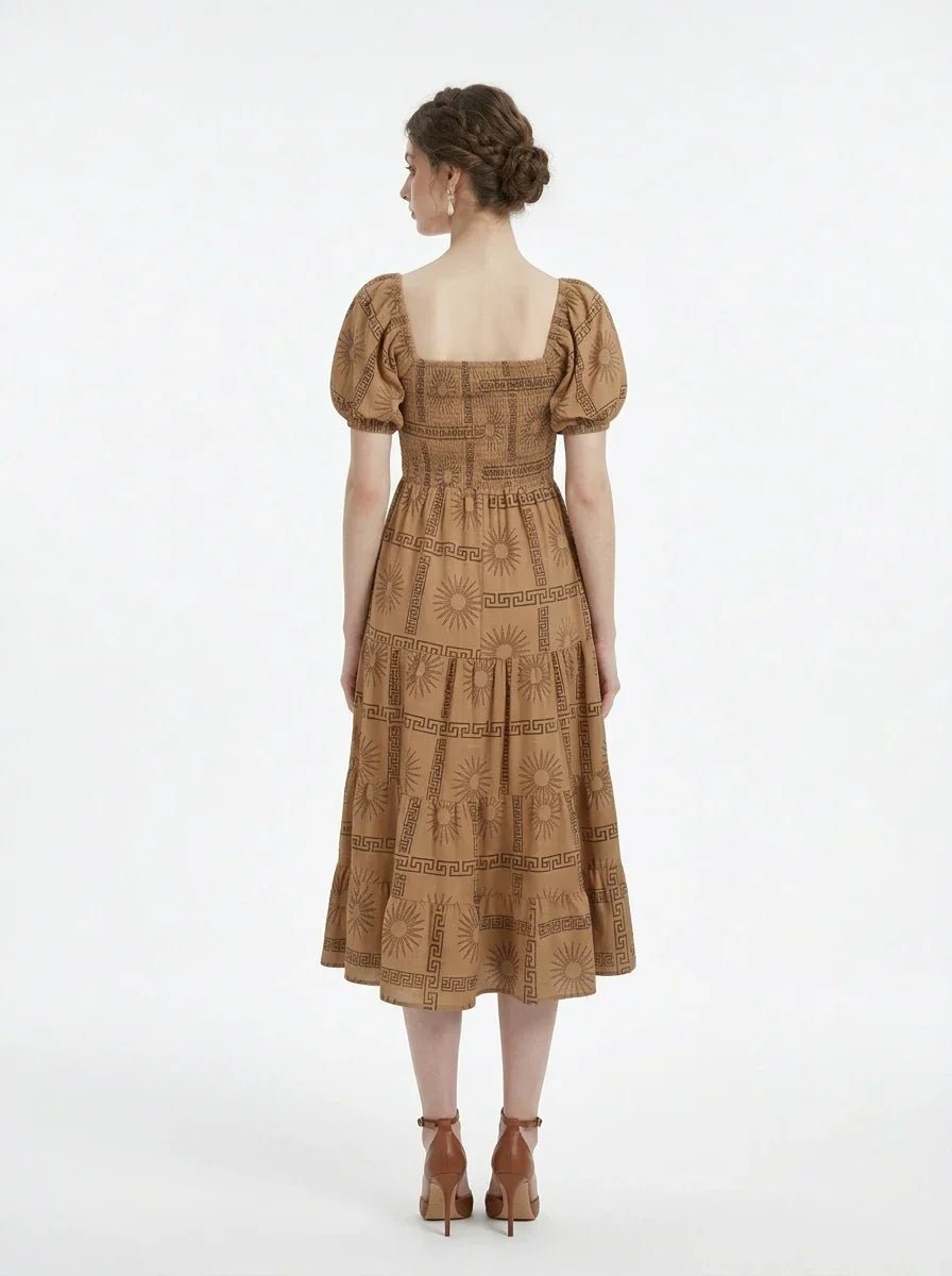Back view of a woman wearing a brown puff-sleeve midi dress with a smocked bodice and tiered skirt featuring a sun and geometric print, standing against a white background.