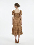 Back view of a woman wearing a brown puff-sleeve midi dress with a smocked bodice and tiered skirt featuring a sun and geometric print, standing against a white background.