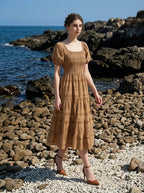 Woman wearing a brown puff-sleeve midi dress with a smocked bodice and tiered skirt featuring a sun and geometric print, walking on a rocky beach with the ocean in the background.