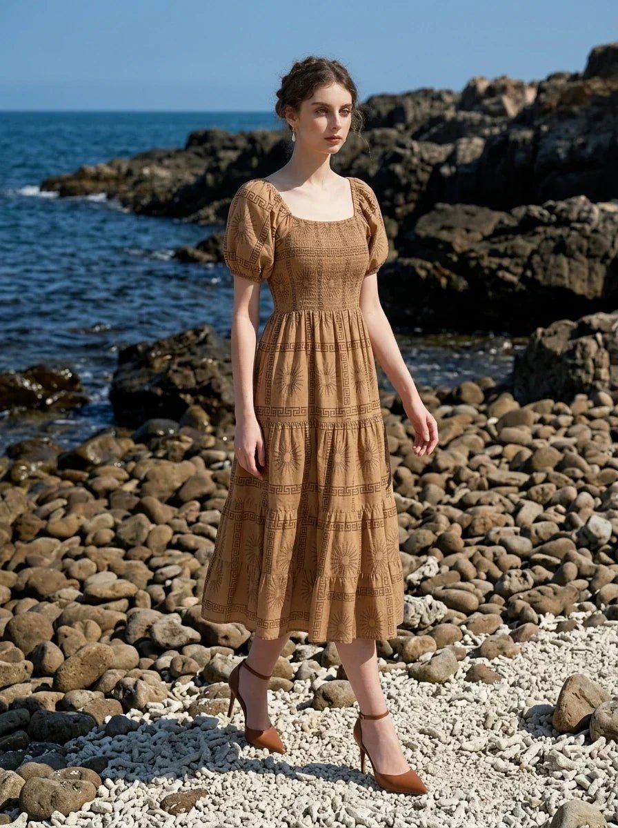 Woman wearing a brown puff-sleeve midi dress with a smocked bodice and tiered skirt featuring a sun and geometric print, walking on a rocky beach with the ocean in the background.