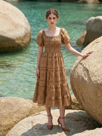 Woman wearing a brown puff-sleeve midi dress with a smocked bodice and tiered skirt featuring a sun and geometric print, leaning on a rock on a beach with the ocean in the background.