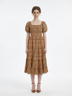 Woman wearing a brown puff-sleeve midi dress with a smocked bodice and tiered skirt featuring a sun and geometric print, standing against a white background.