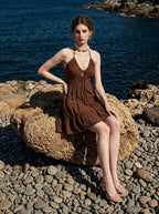 Woman wearing a brown tiered mini halter dress, sitting on a rock on a pebbled beach with the ocean in the background