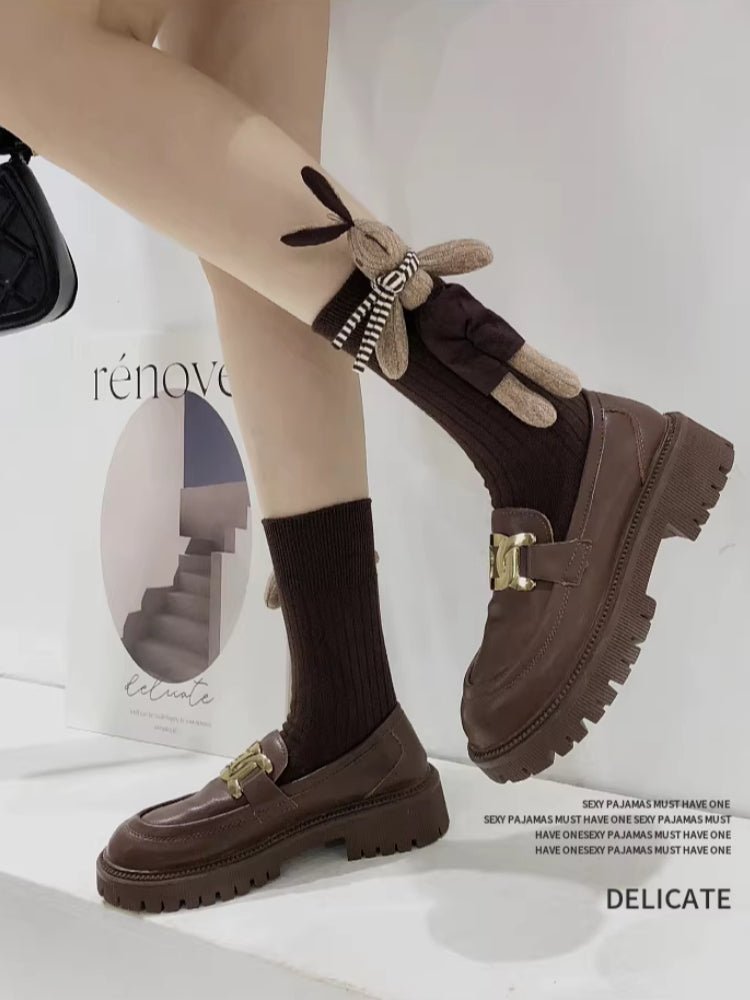 Legs of a person wearing brown knee-high ribbed socks with a plush bunny applique and brown chunky sole loafers.