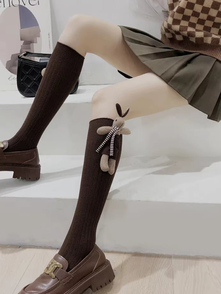 A person sitting, showing their legs in brown knee-high ribbed socks with a plush bunny applique and brown loafers.