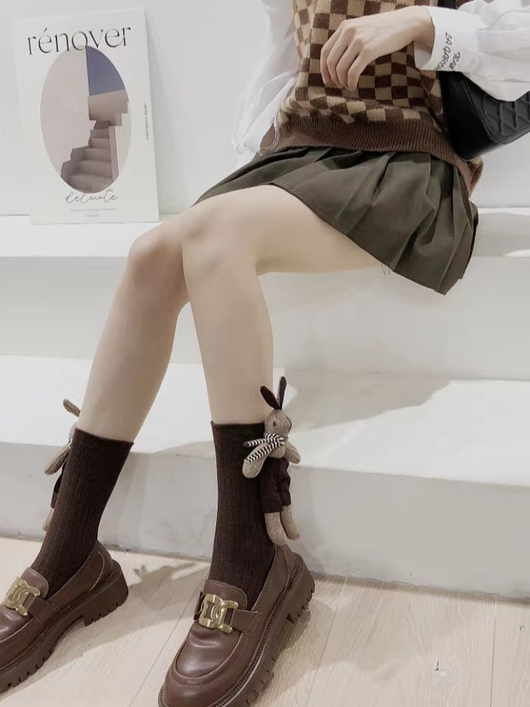 A person sitting down, wearing brown knee-high ribbed socks with a plush bunny applique and brown pleated skirt and loafers.