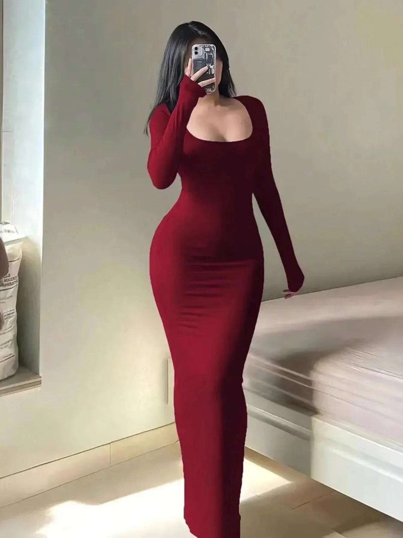 Woman wearing a long-sleeved, scoop-neck, form-fitting, floor-length mermaid silhouette dress in dark red, posing in a room.