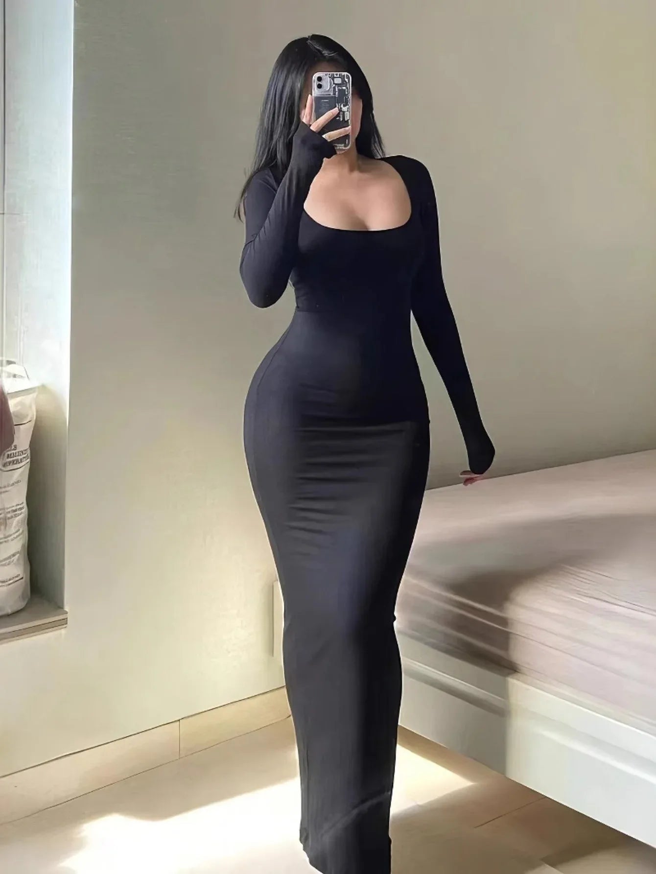 Woman wearing a long-sleeved, scoop-neck, form-fitting, floor-length mermaid silhouette dress in black, posing in a room.