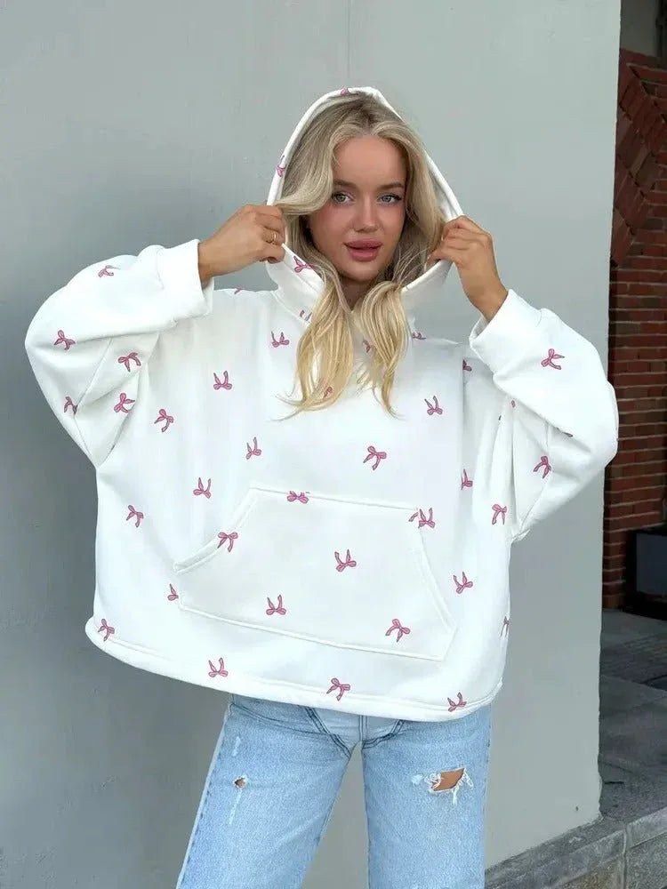 Close-up of a woman wearing an oversized white hooded sweatshirt with a pink bow print, adjusting the hood.
