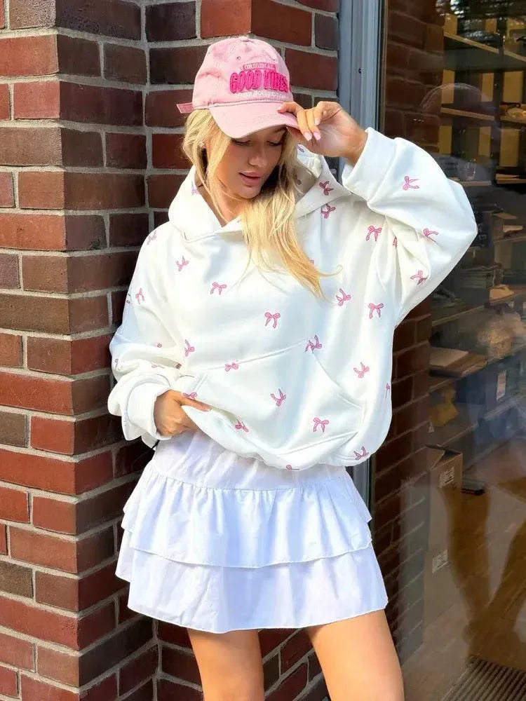 Woman wearing an oversized white hooded sweatshirt with a pink bow print and a white ruffled skirt, accessorized with a pink baseball cap. Main image