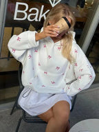 Woman sitting in a chair, wearing an oversized white hooded sweatshirt with a pink bow print and a white ruffled skirt, holding a coffee cup.