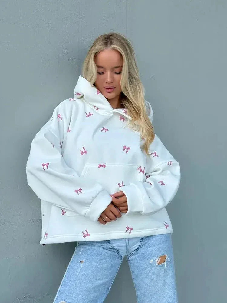 Woman in an oversized white hooded sweatshirt with a pink bow print, paired with distressed light-wash denim jeans.