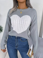 Close-up of a woman wearing a grey knit sweater with a white cable knit heart graphic, blue jeans, and gold necklaces.