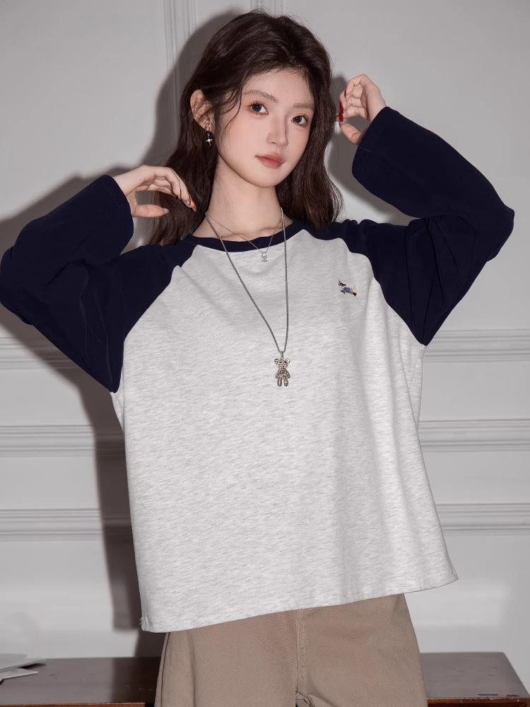 Woman wearing a light grey long-sleeved t-shirt with navy blue raglan sleeves and beige pants, posing with hands near her head.