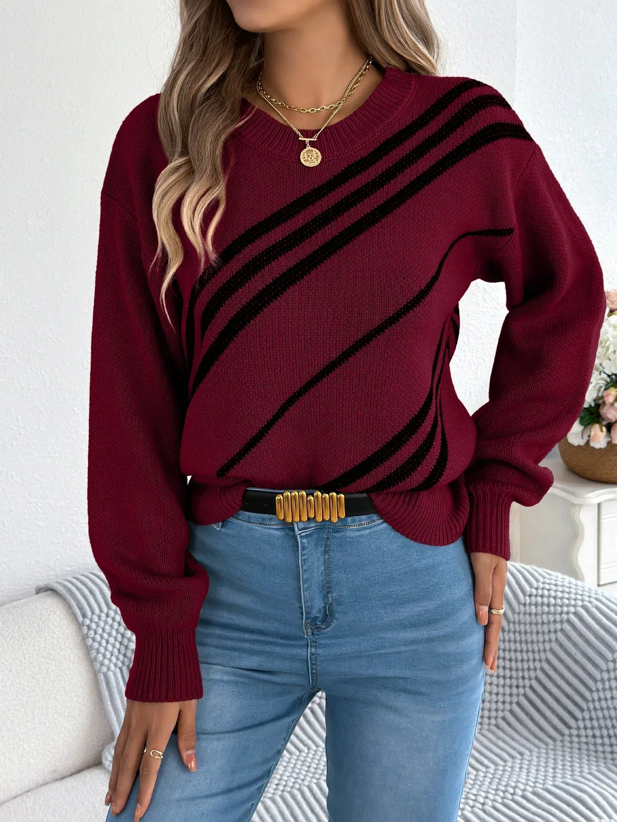 Woman wearing a burgundy knit sweater with black diagonal stripes, blue jeans, and a gold necklace.