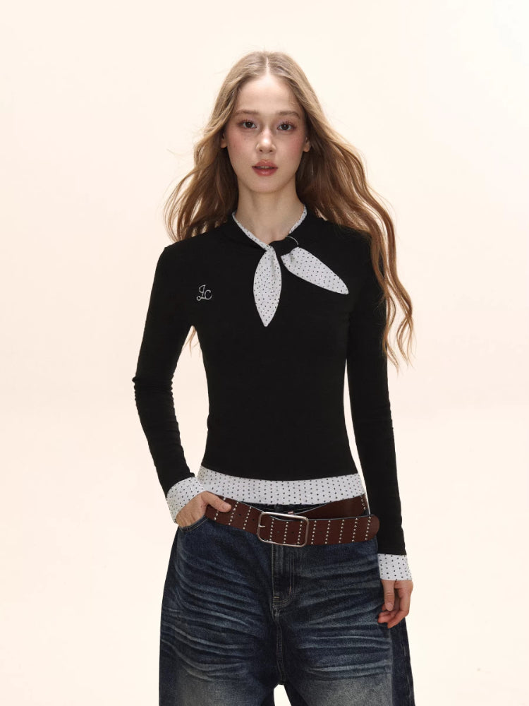 Model from the chest up wearing a black long-sleeve top with a white polka dot scarf detail and white polka dot cuffs, with a small embroidered logo on the chest.