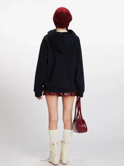 Back view of a woman wearing a dark blue hooded cardigan with colorful fruit buttons, a red beanie, a red faux leather pleated skirt, white knee-high socks, and cream-colored boots.