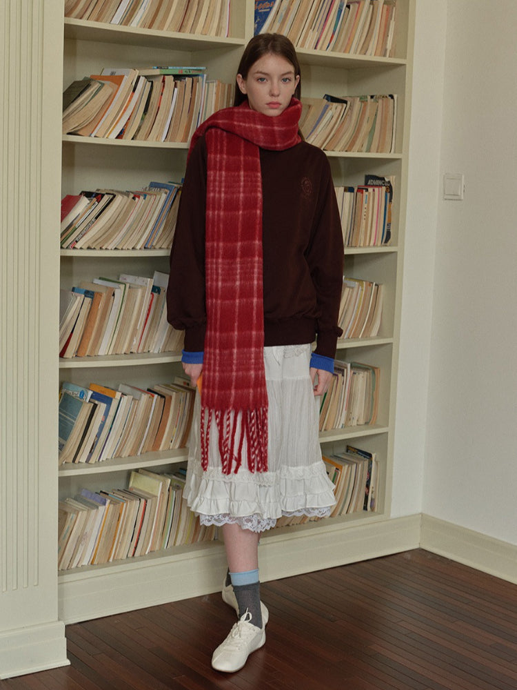 A young woman stands in front of a bookshelf wearing a brown sweatshirt, a white ruffled skirt, and a red plaid scarf.