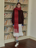 A young woman stands in front of a bookshelf wearing a brown sweatshirt, a white ruffled skirt, and a red plaid scarf.