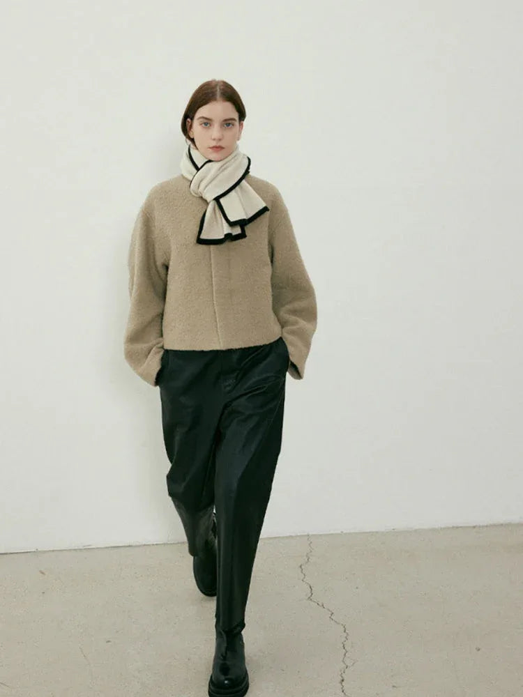 Woman walking towards the camera in a beige textured coat, cream scarf with black trim, and black pants