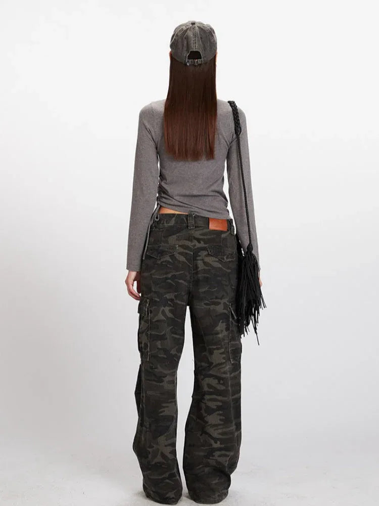 Woman wearing a grey long-sleeved mock neck top with a cropped hem and camouflage print wide-leg cargo pants, accessorized with a grey baseball cap and a black fringe crossbody bag, back view. Secondary image
