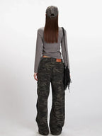 Woman wearing a grey long-sleeved mock neck top with a cropped hem and camouflage print wide-leg cargo pants, accessorized with a grey baseball cap and a black fringe crossbody bag, back view.