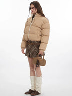 Side view of a model wearing a tan cropped puffer jacket, brown plaid pleated mini skirt, white leg warmers, and brown loafers.