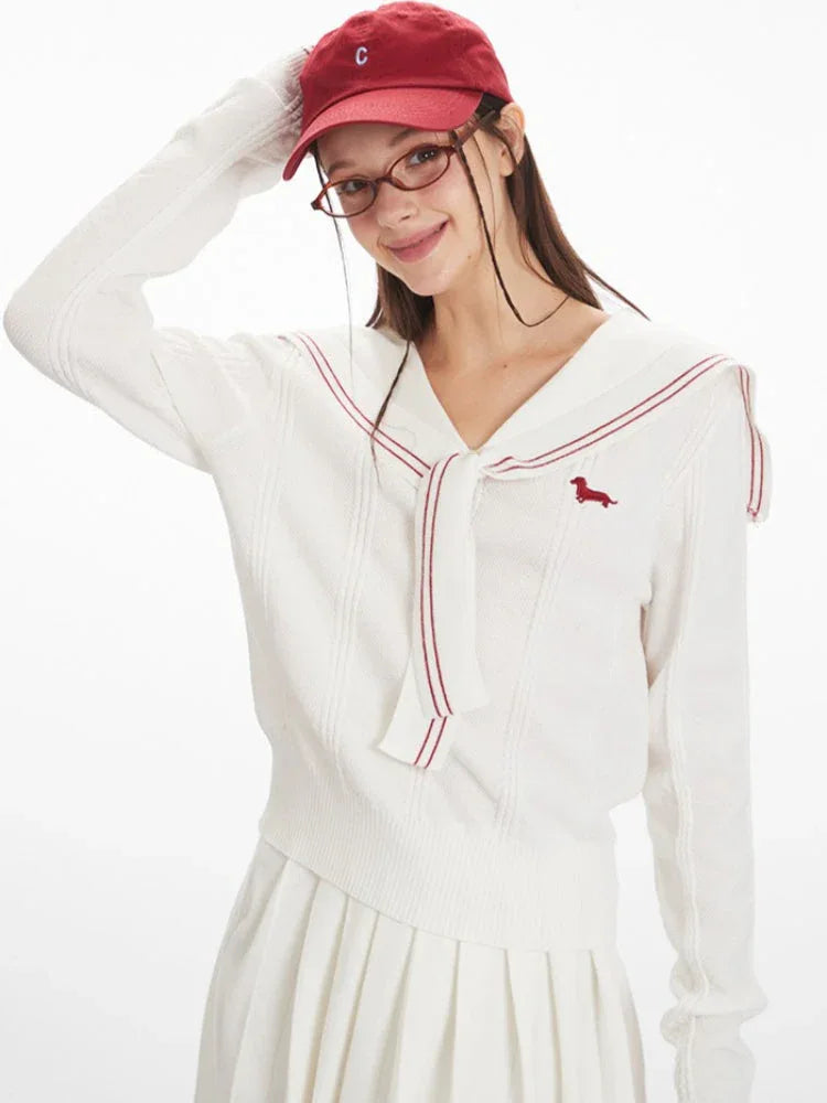 Close-up of a model smiling and touching her hat, wearing a white knit sweater with a sailor collar and red trim.