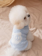 A white bichon frise dog wearing a light blue plush dog sweater with a white 'Wcat' logo patch, sitting on a beige surface, viewed from the side.