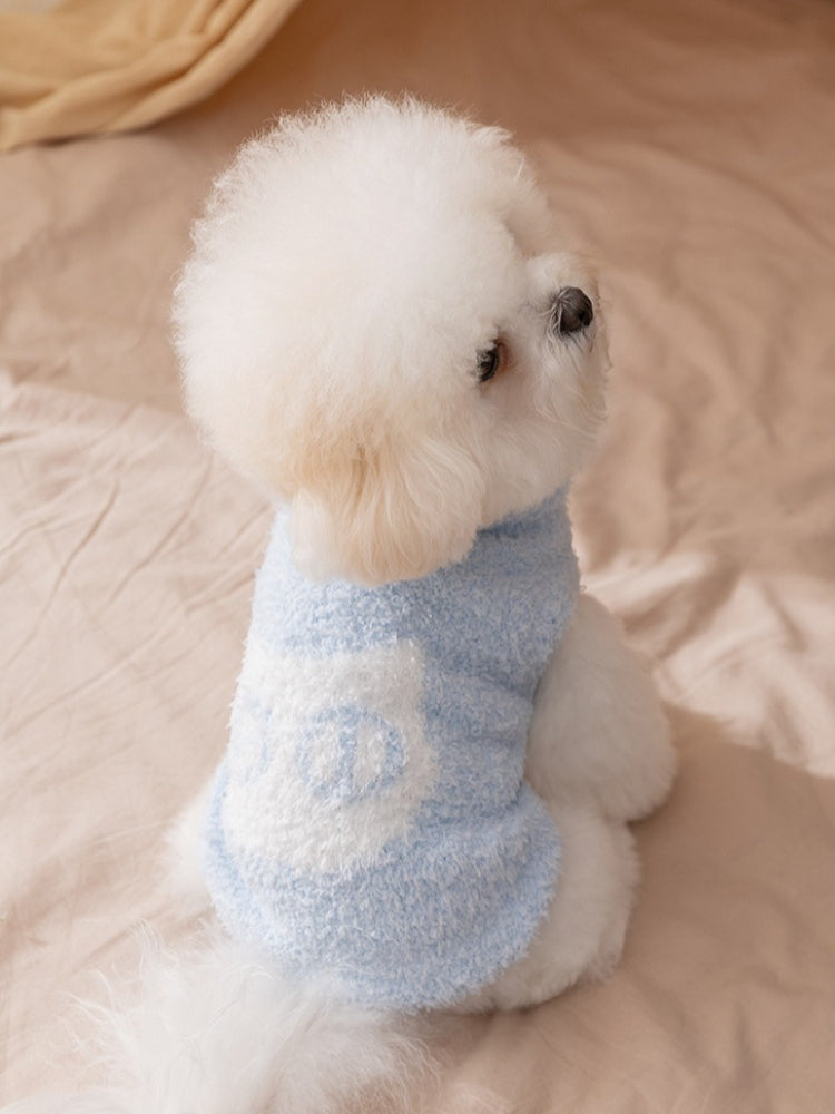 A white bichon frise dog wearing a light blue plush dog sweater with a white 'Wcat' logo patch, sitting on a beige surface, viewed from the side.