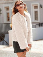 Side view of a woman in a white faux fur cropped jacket, white top, and black shorts, holding a handbag.